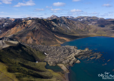 A breathtaking aerial view of a rugged, mountainous landscape featuring a blend of vibrant colors. The scene includes steep slopes with shades of orange, brown, and green, interspersed with patches of snow. The terrain transitions into a calm blue lake that mirrors the sky above, featuring a few rocky outcrops and gentle shorelines. In the background, additional mountain ranges stretch into the distance, showcasing a variety of textures and shapes, under a wide expanse of partly cloudy sky.