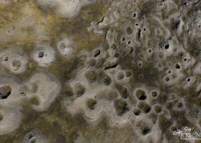 The image features a textured surface resembling a rocky terrain, with numerous depressions and circular shapes scattered across it. The colors vary from gray to earthy tones, with patches of greenish moss or lichen in some areas. The undulating patterns create an organic feel, as if shaped by natural forces over time. The scene gives an impression of a rugged landscape, possibly a volcanic or eroded area, inviting curiosity about its formation and surroundings.