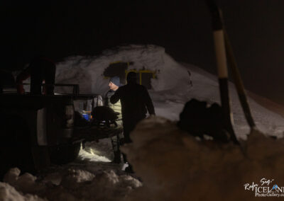 In a dimly lit, snowy environment, two individuals are seen interacting near the back of a vehicle. One person stands on the truck’s edge, possibly retrieving or organizing equipment, while the other, wearing a dark sweater and a beanie, is engaged in conversation or observation. The area is heavily blanketed in snow, with shadows cast by the sparse illumination. In the background, a sizable mound of snow looms, hinting at a remote, wintery location. The atmosphere feels cold and slightly mysterious, emphasizing the isolation of the scene.