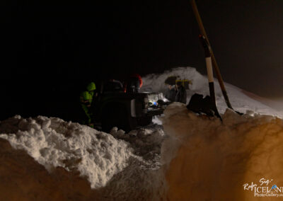 The image depicts a nighttime scene in a snowy landscape. A pile of snow is prominently featured in the foreground, creating a textured white mass. In the background, there are three individuals working near an off-road vehicle, which appears to be parked on a snow-covered path. One person, dressed in a bright green jacket, is leaning against the vehicle, while another person is standing by its side, and a third is climbing onto the vehicle. The surrounding area is dimly lit, with a slight illumination from the vehicle, indicating a desolate but active scene in a winter environment. Snow covers the ground extensively, creating a sense of isolation and adventure.