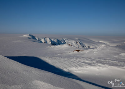 A vast landscape covered in thick, undisturbed snow stretches across the scene, reflecting bright sunlight. The snow forms gentle, rolling hills and mounds, creating subtle curves and shadows. In the distance, rugged mountains tower above the snowy expanse, their peaks slightly visible. The sky above is clear and bright blue, enhancing the stark contrast between the white snow and the sky. The overall atmosphere conveys a sense of tranquility and pristine wilderness.