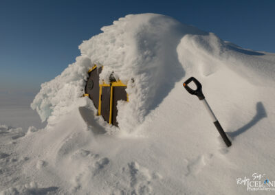 A small, snow-covered structure is partially visible, with the roof heavily coated in white snow and ice. The walls of the building are dark brown, and there are bright yellow accents around a door that is almost completely buried. A black and white snow shovel stands in the snow nearby. The background features a clear blue sky, contrasting with the white landscape, and there are gentle snowdrifts surrounding the building, suggesting a cold, wintry environment.