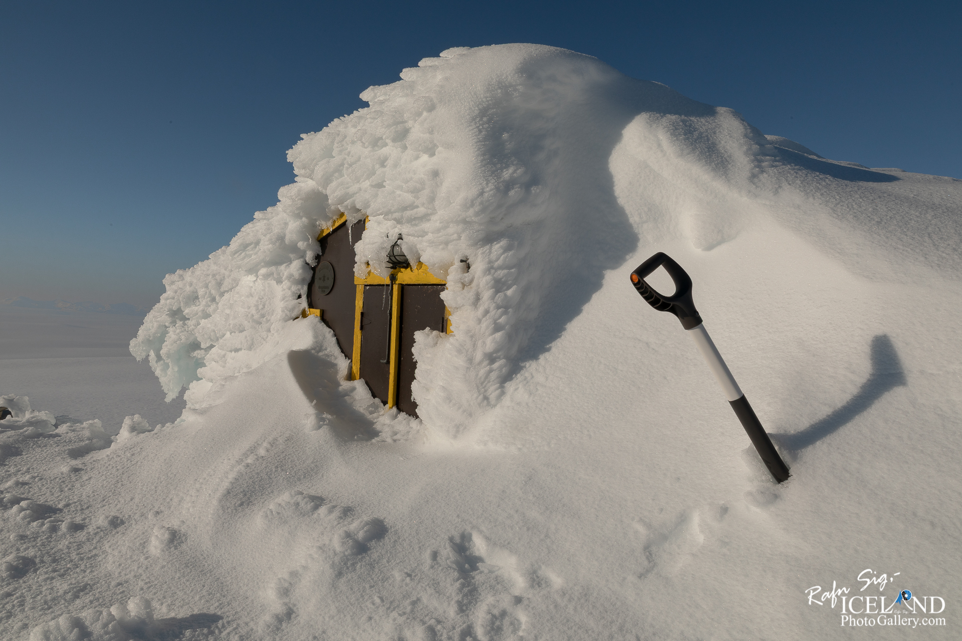 A small, snow-covered structure is partially visible, with the roof heavily coated in white snow and ice. The walls of the building are dark brown, and there are bright yellow accents around a door that is almost completely buried. A black and white snow shovel stands in the snow nearby. The background features a clear blue sky, contrasting with the white landscape, and there are gentle snowdrifts surrounding the building, suggesting a cold, wintry environment.