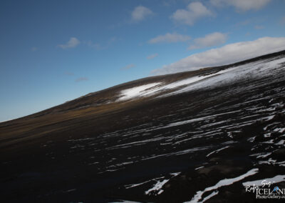 A view of a sloping volcanic landscape under a mostly clear blue sky. The ground is predominantly dark, consisting of volcanic rock and ash, with streaks of white snow visible across the surface. The hill rises gently in the distance, showcasing a mix of earthy tones, with some patches of grass peeking through. Wispy clouds drift lazily in the sky, enhancing the serene and rugged nature of the scene.