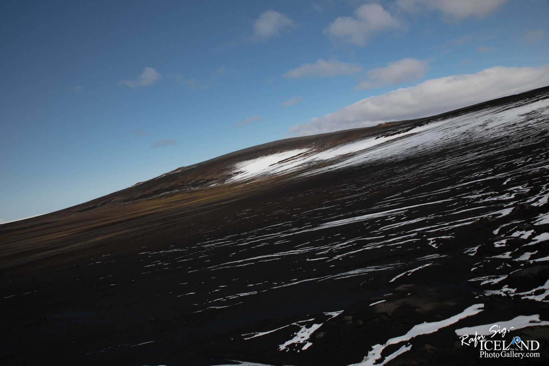 A view of a sloping volcanic landscape under a mostly clear blue sky. The ground is predominantly dark, consisting of volcanic rock and ash, with streaks of white snow visible across the surface. The hill rises gently in the distance, showcasing a mix of earthy tones, with some patches of grass peeking through. Wispy clouds drift lazily in the sky, enhancing the serene and rugged nature of the scene.