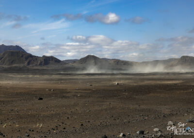 A vast, barren landscape stretches across the scene, characterized by a mix of dark earth and scattered rocky formations. In the background, rugged mountains rise against a sky filled with patches of soft clouds. The ground is mostly dry and cracked, with only a few patches of green grass peeking through the dust. A light wind appears to lift dust particles into the air, creating small clouds that drift across the flat terrain. The overall atmosphere gives a sense of desolation and raw natural beauty.