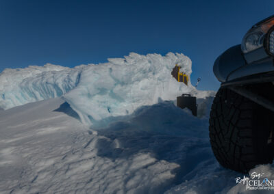 A close-up view of a snow-covered landscape under a clear blue sky. In the foreground, the rugged tread of a black vehicle tire is visible, suggesting it is parked on deep snow. Behind the tire, large snowdrifts rise up, featuring jagged edges and icy textures that hint at their recent formation. In the background, a structure is partially obscured by the snow, with a yellow element peeking through and a shovel resting nearby, indicating the snowy environment's utility. The scene conveys a sense of winter wilderness and exploration.