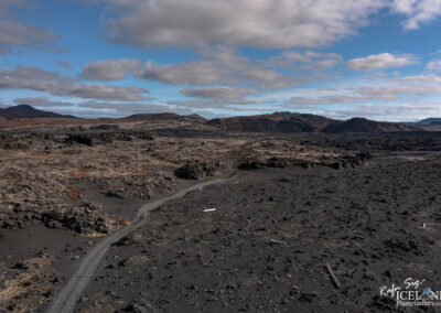 The image shows a rugged, expansive volcanic landscape characterized by dark, rocky terrain scattered with patches of lighter stones. A winding dirt path traverses through the scene, leading towards distant mountains under a sky filled with soft, fluffy clouds. The overall atmosphere is stark and barren, with an otherworldly feel, highlighting the raw beauty of nature shaped by volcanic activity.