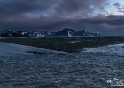 A tranquil landscape scene depicting a rocky terrain at the foot of prominent, snow-capped mountains under a cloudy sky. The foreground features a winding waterway flowing gently, surrounded by pebbles and stones. In the background, dramatic peaks rise sharply, partially obscured by low-hanging clouds, creating a moody atmosphere. The color palette consists of muted blues and grays, evoking a sense of calm and solitude in this remote natural setting.