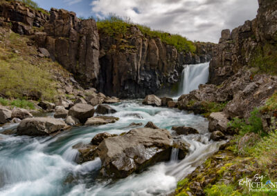 The image showcases a scenic natural landscape featuring a waterfall cascading down rocky cliffs. The waterfall is framed by jagged, dark rocks that rise prominently from the ground. At the base of the waterfall, a river flows swiftly, with clear, turquoise water swirling among large boulders and smaller rocks scattered along the riverbanks. The surrounding terrain is lush with green vegetation, contrasting with the rocky surfaces. Above, the sky is partially cloudy, allowing some sunlight to illuminate the scene, enhancing the vibrant colors and textures of the landscape.