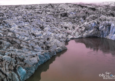 The image showcases a vast glacier with a textured, jagged surface that features a mix of icy blues and whites, along with darker streaks indicating areas of dirt or rock. The glacier meets a body of water that reflects subtle hues, giving it an amber-pinkish appearance. The landscape is expansive, with the glacier extending into the distance, under a cloudy sky that adds a sense of moody ambiance to the scene. The overall setting evokes a feeling of coldness and isolation, typical of a remote icy environment.