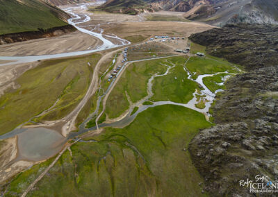 Aerial view of a rugged landscape featuring winding rivers and streams that cut through a patchwork of green and brown earth. There are several small clusters of vehicles parked near a campsite, surrounded by grassy areas and rocky hills in the distance. The terrain is uneven, with a mix of rocky outcrops and smooth paths. The scene suggests a remote and natural setting, likely in a mountainous region. The colors range from deep greens of vegetation to earthy browns and grays of the soil and rocks.