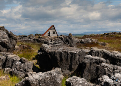 A picturesque scene showcasing a small, A-frame wooden house with a red roof, set against a rugged landscape of dark, rocky formations. The house has large windows, allowing light to shine in. Surrounding the house, there are tufts of green grass and patches of earth, contrasting with the stark rocks. In the background, rolling hills and mountains rise beneath a partly cloudy sky, adding depth to the scenery. This tranquil setting evokes a sense of isolation and natural beauty, characteristic of a remote outdoor environment.