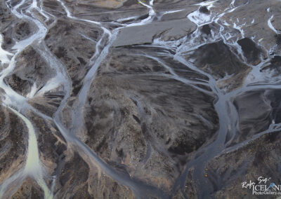 The image features a bird's-eye view of a unique landscape characterized by dark, textured earth and meandering streams. The ground has a mostly dark grayish-black hue, with patches of lighter colors and a glossy sheen where water flows. The streams trace intricate, winding paths through the terrain, creating a natural network that is both organic and rhythmic. The area appears rugged and barren, with scattered stones and gravel adding texture to the surface. The overall scene evokes a sense of vastness and raw beauty in nature.