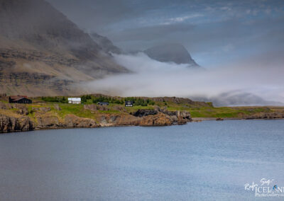 A serene coastal landscape featuring a calm body of water reflecting the sky. In the foreground, rocky shorelines blend into green grassy areas. Scattered across the landscape are a few small houses, including one with a red roof and others in white and dark colors, nestled against the backdrop of towering mountains. The mountains are partially obscured by low-hanging clouds, creating a mystical atmosphere. The scene is peaceful, with an air of solitude and natural beauty.